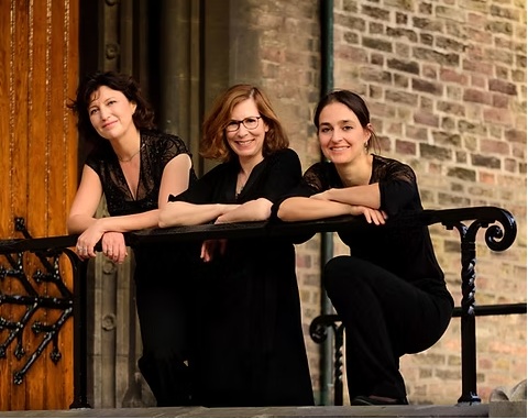 Drei schwarz gekleidete Frauen lehnen über einem Geländer vor einem historischen Backsteingebäude